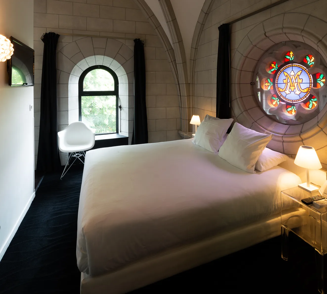 Hotel room with a large bed, stained-glass window, and arched stone walls.