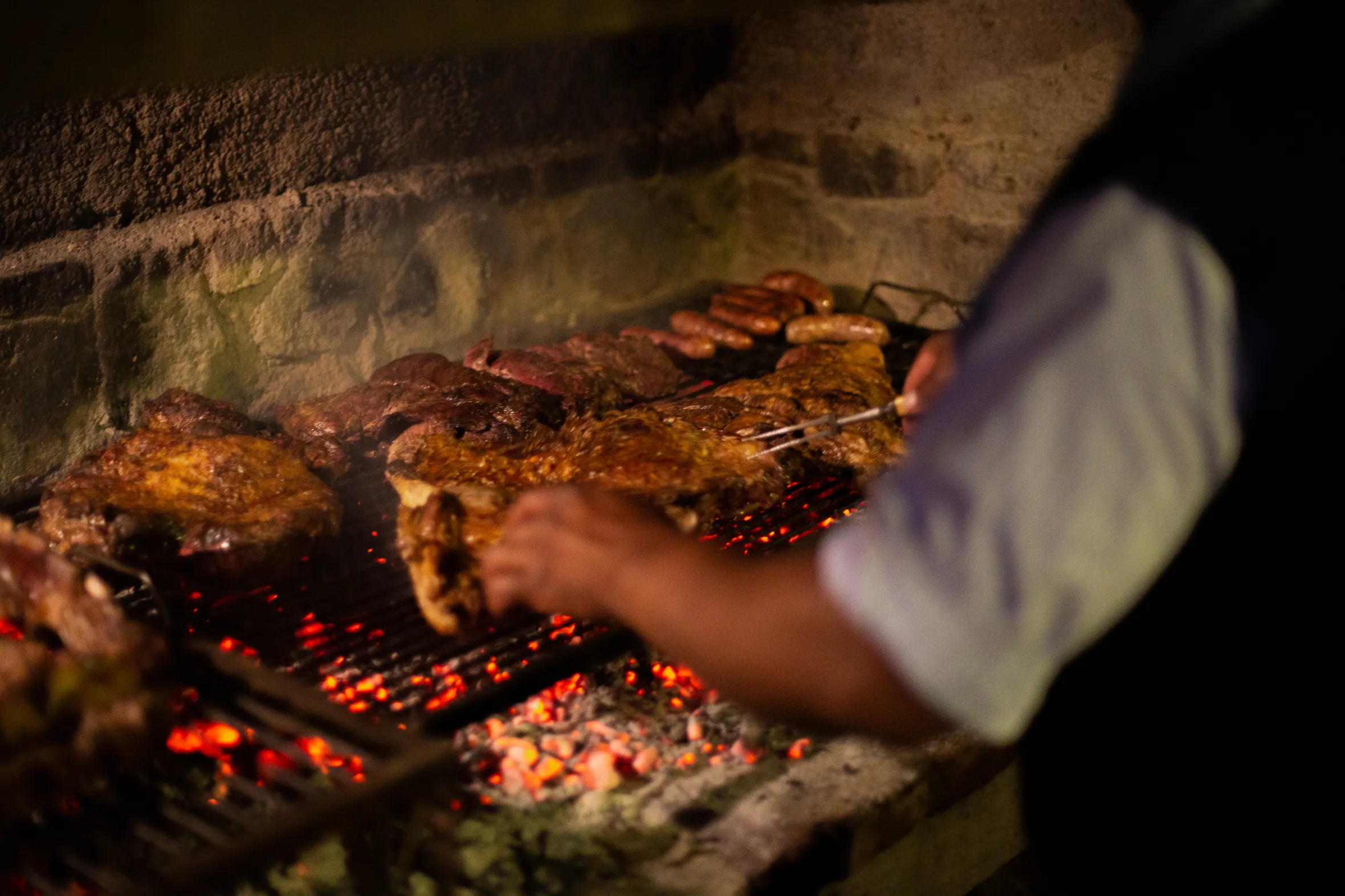 Meat and sausages cooking on a grill over red hot coals, with a person turning the meat.