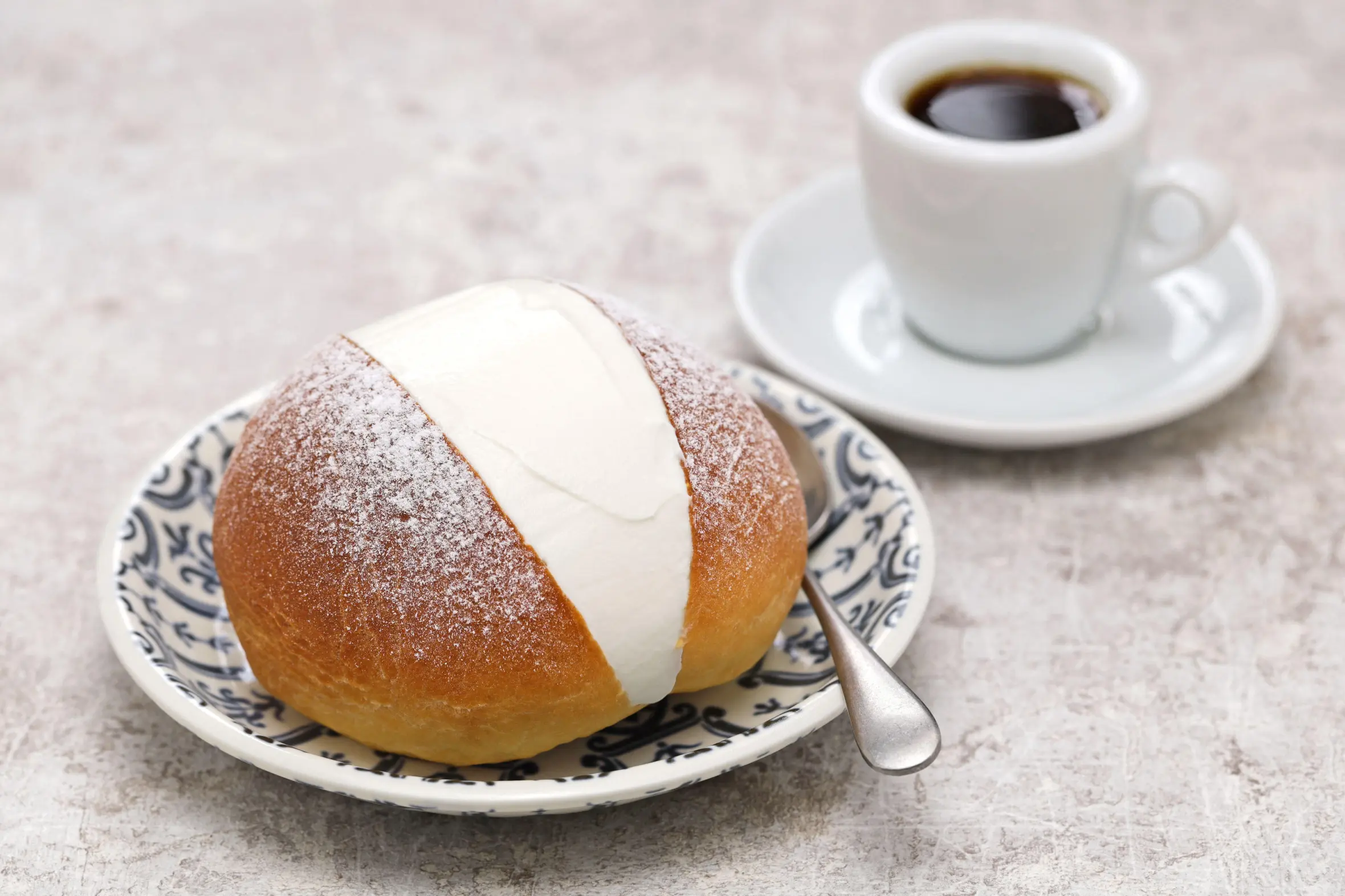 A Maritozzo sweet bun filled with whipped cream and dusted with powdered sugar, served on a patterned plate with a spoon, next to a cup of espresso.