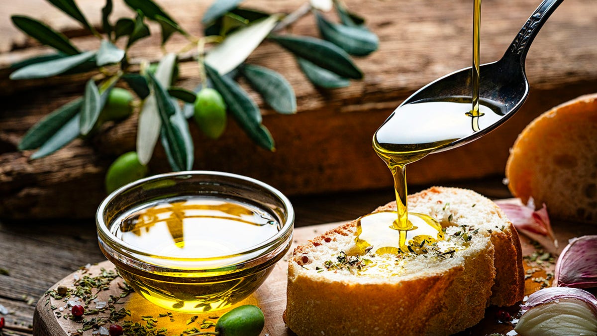 Extra virgin olive oil pouring from spoon to a bread slice on rustic wooden table.