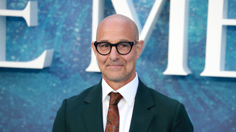 Stanley Tucci, from the shoulders up, in a suit, tie, and glasses