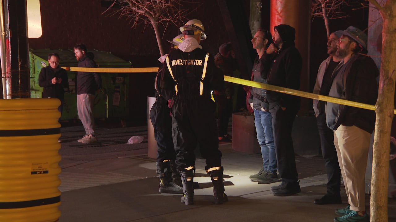 Firefighters with the words 'investigator' on their backs are seen near fire tape, with other people milling around.