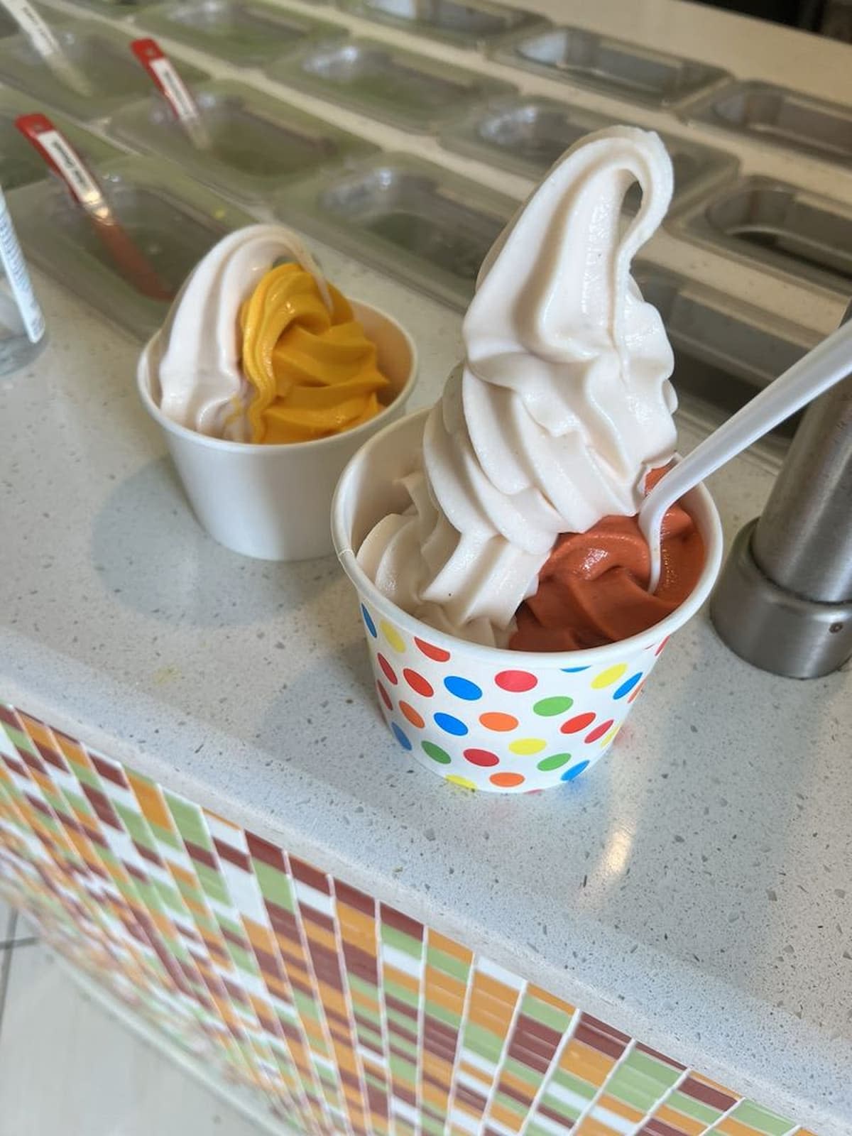 Two cups of soft serve frozen yogurt sit on a counter. One cup has vanilla and orange flavors with a spoon, the other has vanilla and a yellow flavor. Colorful mosaic tiles decorate the counter’s side.