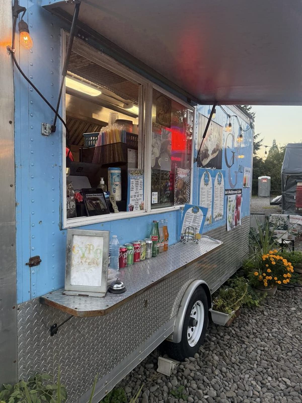 A blue and silver food truck with an open service window displays various drinks, menus, and condiments. Decorative lights hang above, with flowers and gravel visible in the foreground.