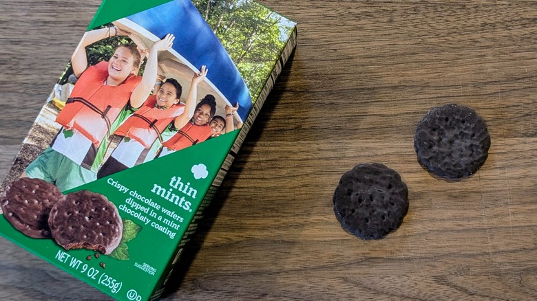 A box of Thin Mints next to two cookies on a wood board