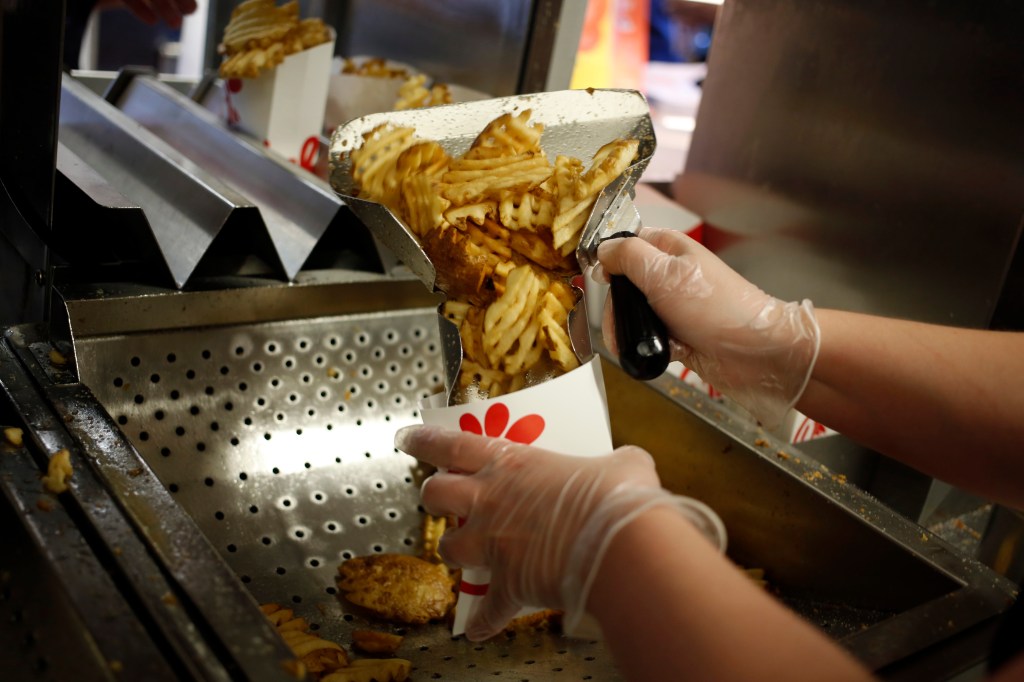 An employee wearing clear gloves prepares waffle fries in a white paper container at a Chick-fil-A restaurant.