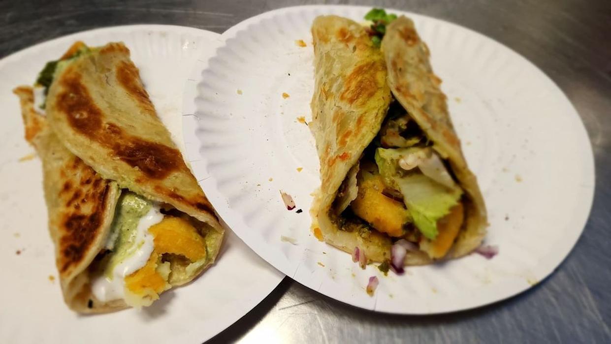 Two stuffed parathas or wraps filled with vegetables, sauce, and possibly paneer, served on white paper plates on a metal surface. One wrap is open at the end, showing colorful fillings.