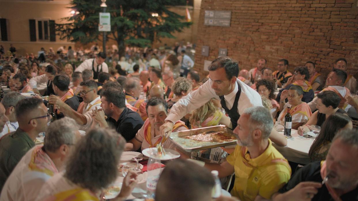 A waiter serves hot food from a tray to a participant at a bustling Palio dinner, held on the lively streets of Siena on the eve of the famed race.