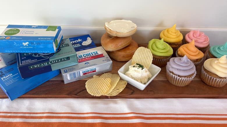 cream cheeses on a wooden cutting board with bagels, cupcakes and chips