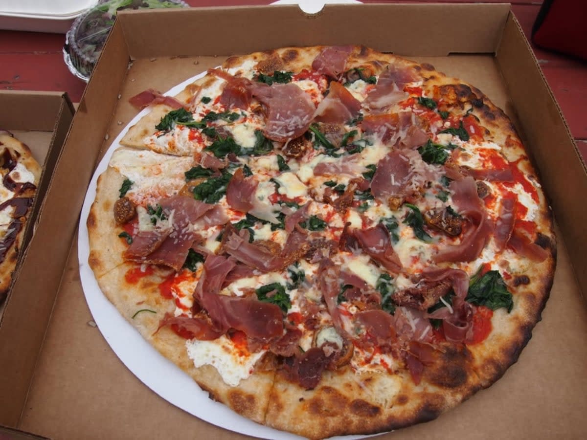 A freshly baked pizza in an open cardboard box, topped with prosciutto, spinach, melted cheese, tomato sauce, and a crispy, slightly charred crust.