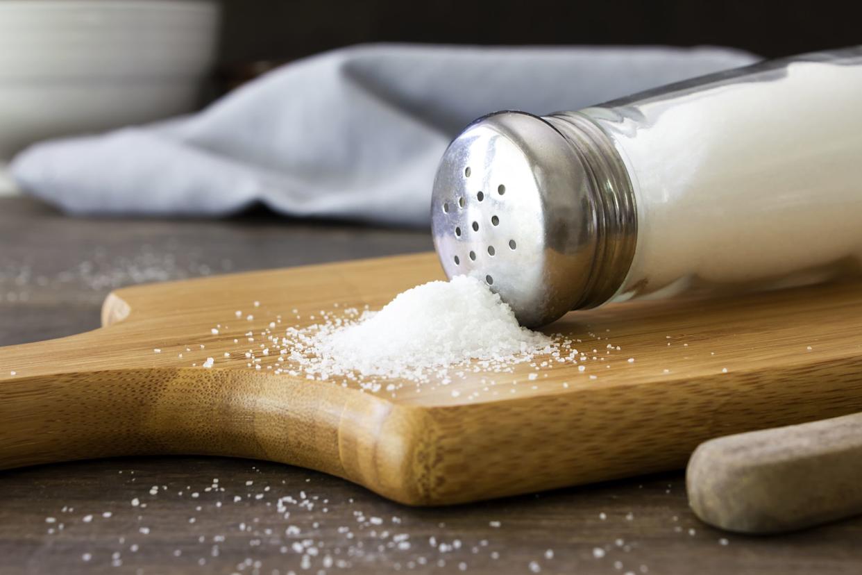 A close up shot of salt spilling out of a salt shaker on to a wooden chopping board. Shot on a rustic wooden kitchen table with kitchen utensils in the background.
