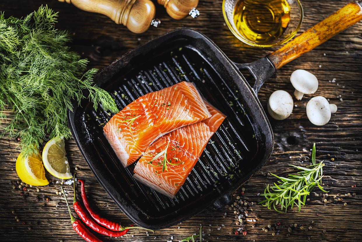 Two raw salmon fillets ready to be grilled in a black cast iron skillet. Various ingredients, such as fresh dill and rosemary, lemon slices, chili peppers, mushrooms, and olive oil, are arranged around the skillet on a dark wooden surface, suggesting the preparation of the dish.