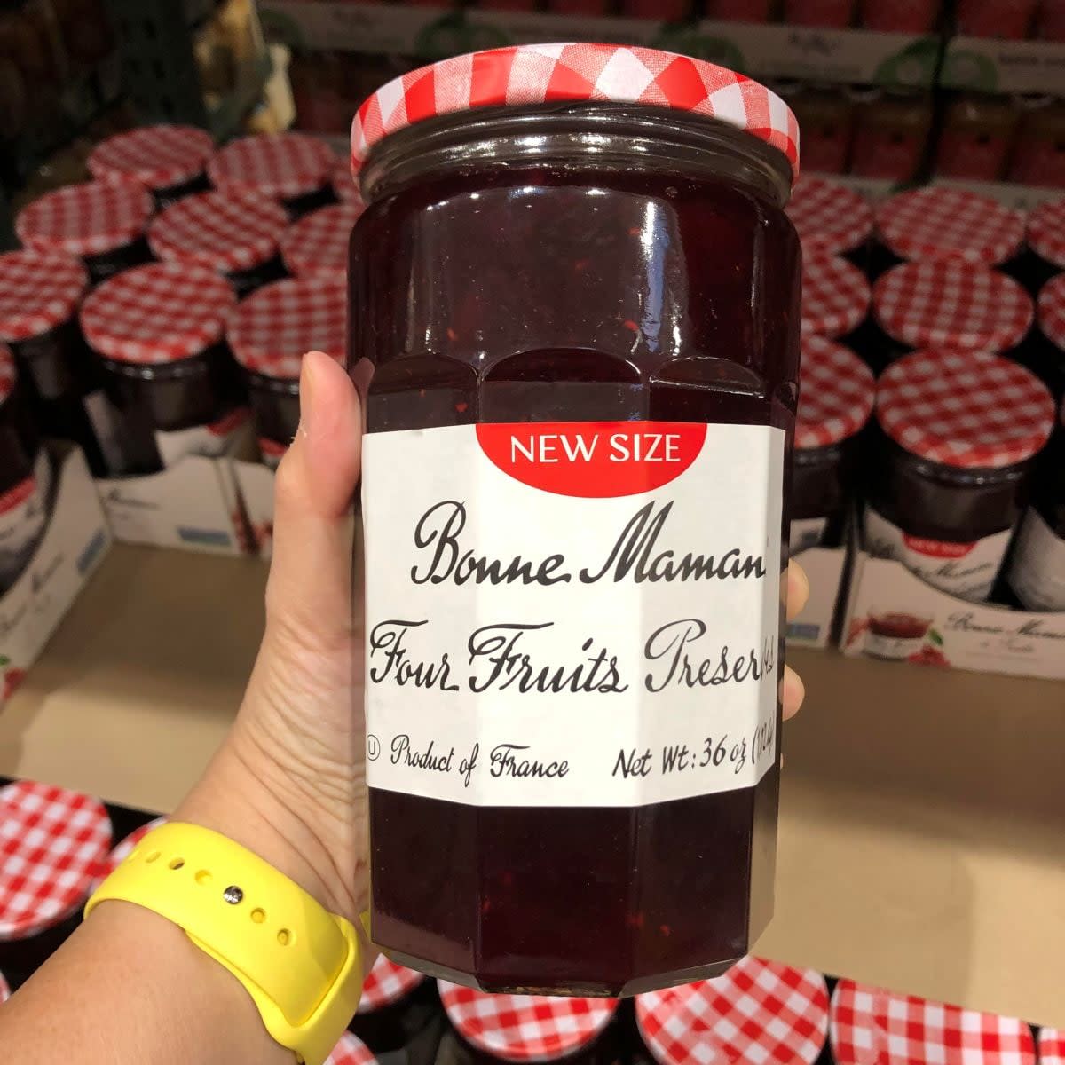 A hand wearing a yellow watch holds a jar of Bonne Maman Four Fruits Preserves with a red and white checkered lid in a store, with similar jars on the shelf in the background.
