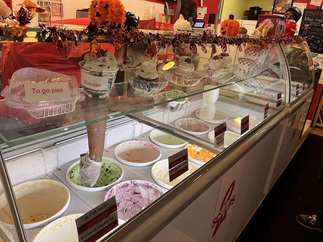 An ice cream shop display case with various ice cream tubs, cones, and toppings. Labels show flavors like butter pecan and banana cream pie. Halloween decorations with orange garland hang above the counter.