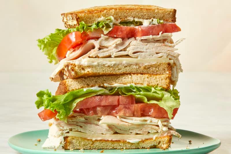 Head on view of a stacked turkey sandwich with cheese, turkey, tomato and lettuce on white bread and a blue plate.
