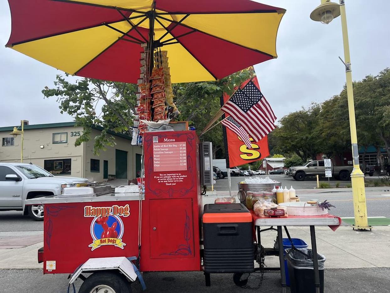 A red hot dog cart with a large yellow and red umbrella, American flag, condiments, and a menu stands on a sidewalk near a street and buildings. Sausages are displayed on skewers above the cart.