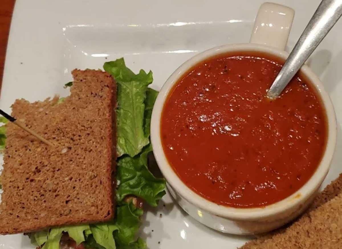 A white cup filled with soup and a spoon sits next to a sandwich with lettuce on whole grain bread, served on a white plate—comfort food that could rival the best soup in every state.