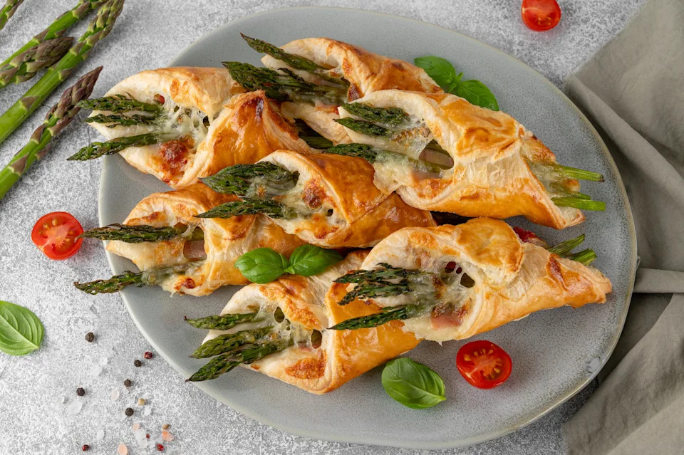 Baked puff pastry with green asparagus, prosciutto or ham and cheese on a gray concrete background. Top view
