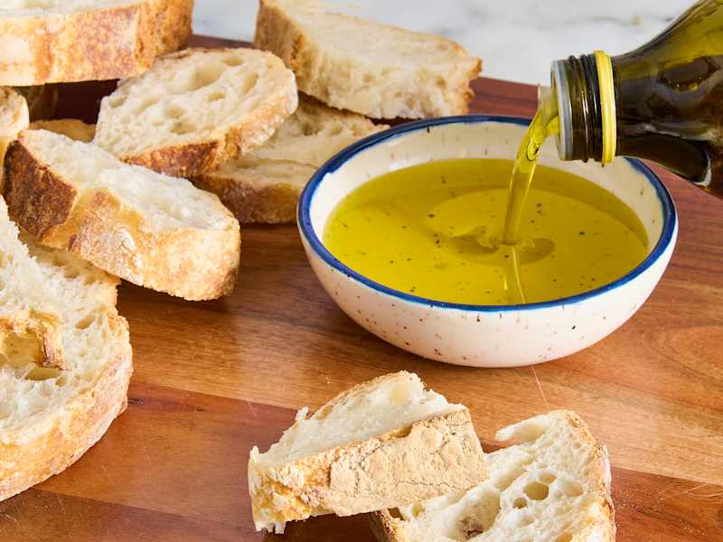 Sliced bread on a wooden board with a bowl of olive oil being poured into it.