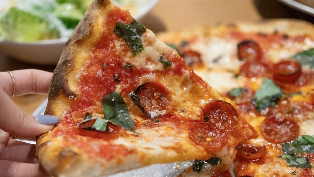 A hand holds a slice of pizza topped with pepperoni, fresh basil, tomato sauce, and melted cheese. The pizza sits on a plate, with a bowl of salad visible in the background.