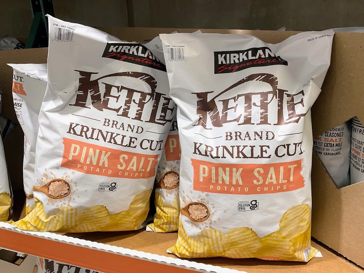 Bags of Kirkland Signature Kettle Brand Krinkle Cut Pink Salt Potato Chips are displayed on a store shelf, with some bags arranged upright and others lying on their side.
