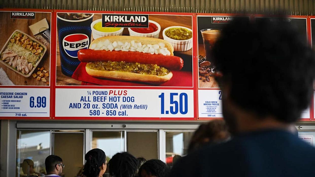 A person stands in line facing a Costco food court menu featuring the $1.50 hot dog and soda combo—one of the classic Costco products loved by shoppers—plus images of other food options and prices above the counter.