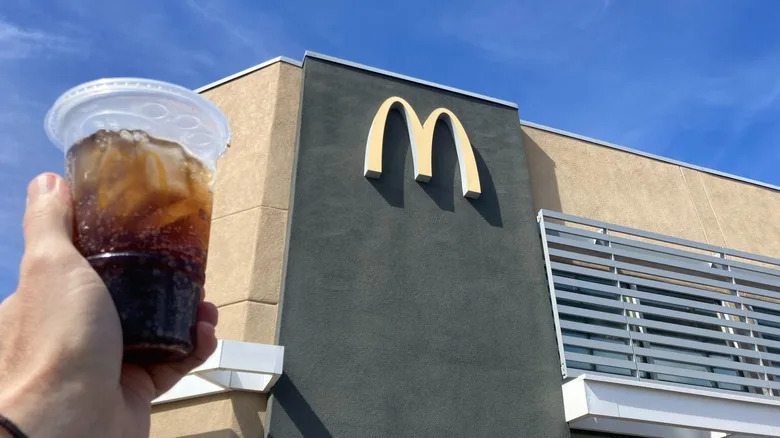 Hand holding a plastic cup of Coke in front of McDonald's storefront during daytime