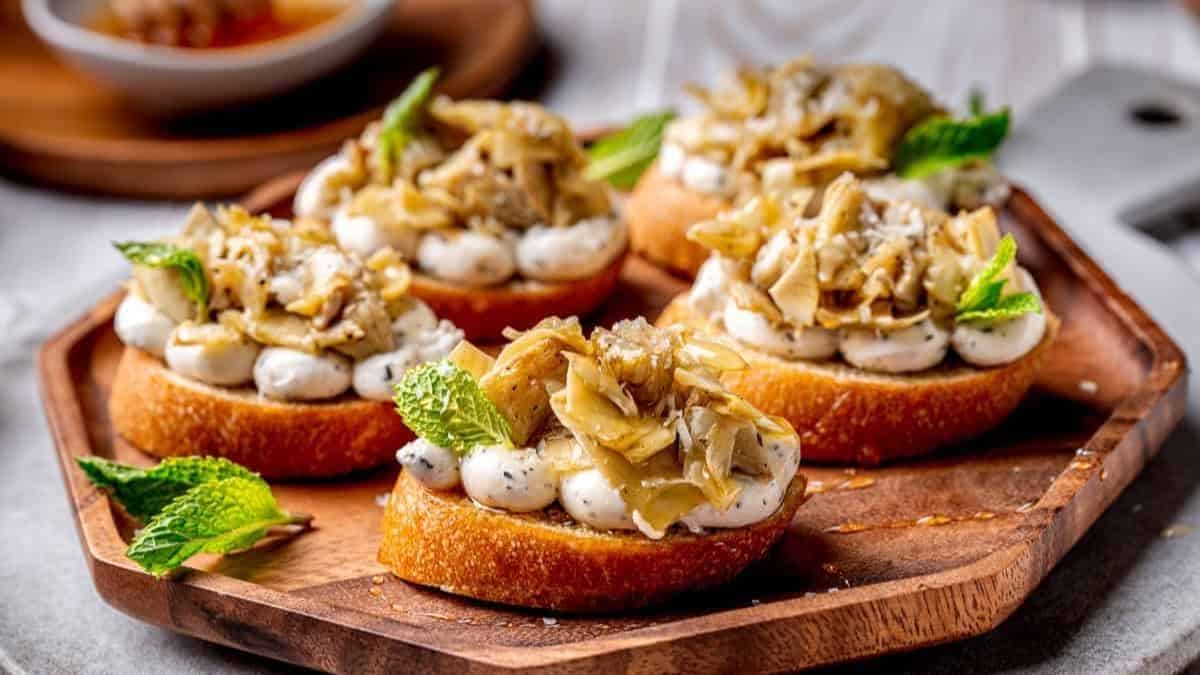 A wooden plate with four pieces of Crostini topped with Marinated Artichokes.