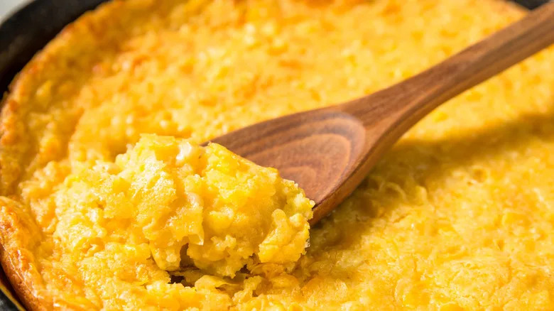 A close up for corn pudding being spooned out of a cast iron skillet with a wooden spoon