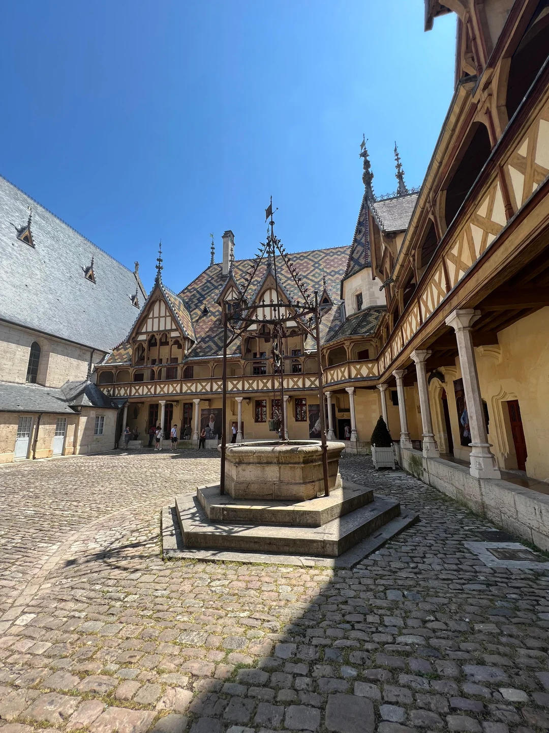 A cobblestone courtyard with a central stone well, surrounded by historic buildings with patterned tile roofs, decorative woodwork, and arched walkways under a bright blue sky.