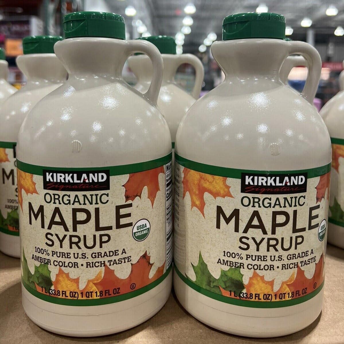 Two large bottles of Kirkland Signature Organic Maple Syrup, one of the Costco products loved by shoppers, are displayed side by side on a store shelf. The beige bottles with green caps feature a colorful maple leaf label.
