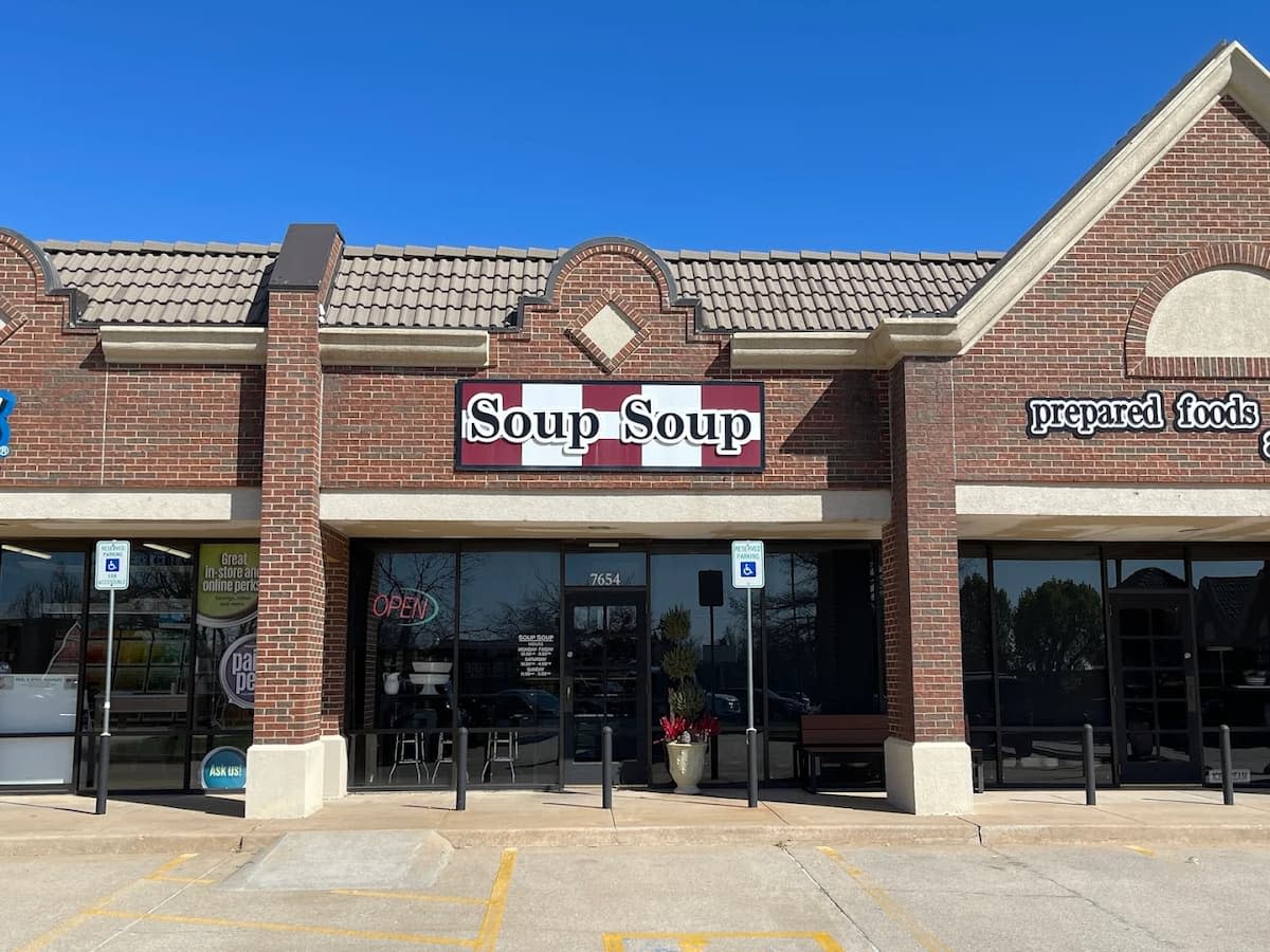 A brick strip mall storefront with a sign reading "Soup Soup" above the entrance. Glass doors and windows reveal a cozy spot to try the best soup in every state. A couple of chairs sit outside, with accessible parking in front and a clear blue sky above.