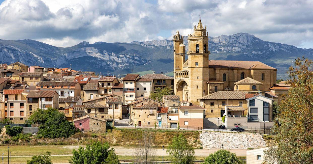 A picturesque Spanish village with a large stone church, surrounded by rustic houses and set against a backdrop of rugged mountains under a partly cloudy sky.