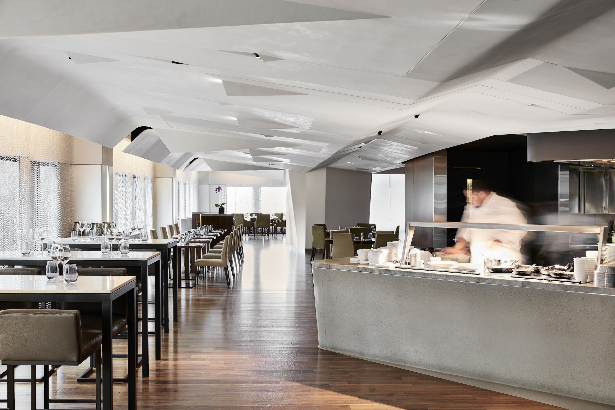 Spacious, modern restaurant dining room with geometric white ceiling, wood floors, rows of neatly set tables, and a chef working at an open kitchen counter.