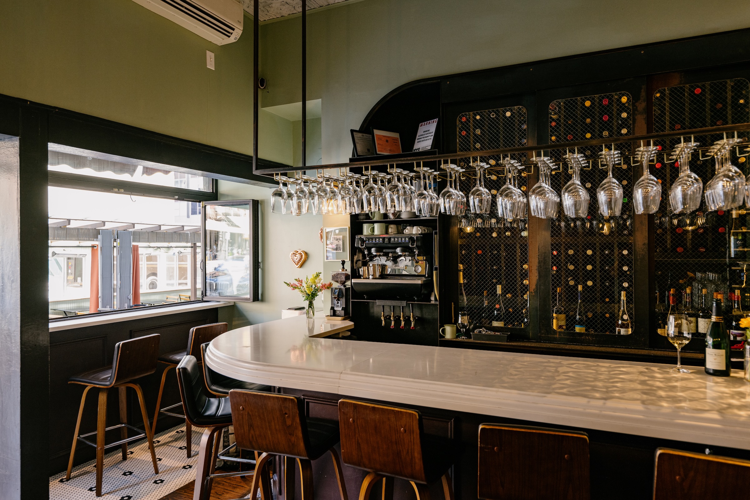 Cozy wine bar with a curved white countertop, wooden stools, wine glasses hanging overhead, and a wall of wine bottles behind the bar.