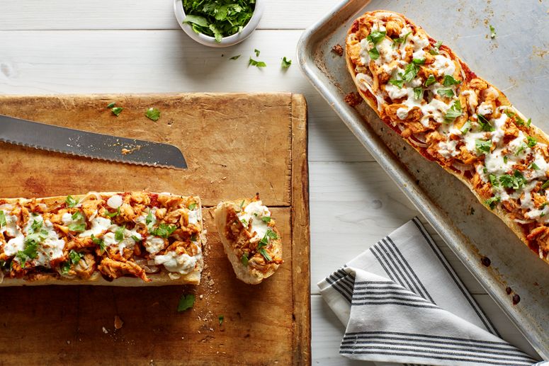 Two loaves of French bread pizza with shredded chicken and mozzarella cheese one on a cutting board and one on a sheet pan.