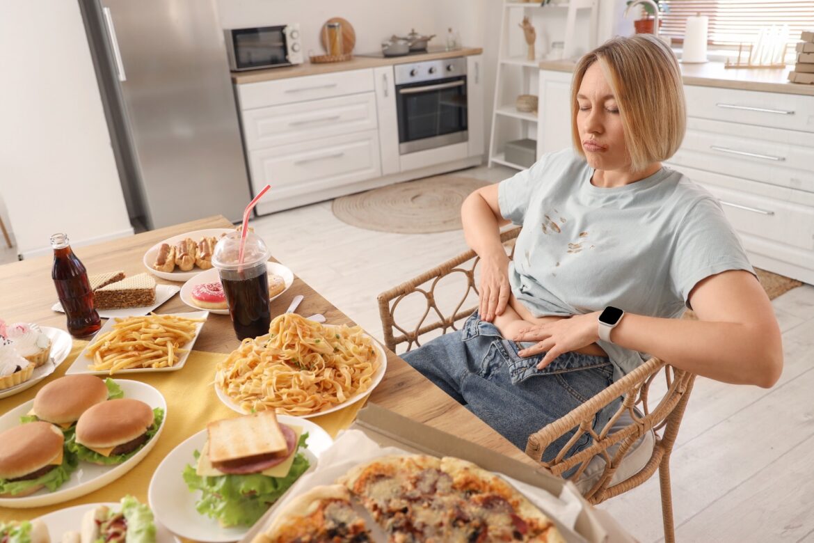 Woman sat at a dining table with lots of food on. Food on her shirt and her jeans are unfastened to show she has overeaten
