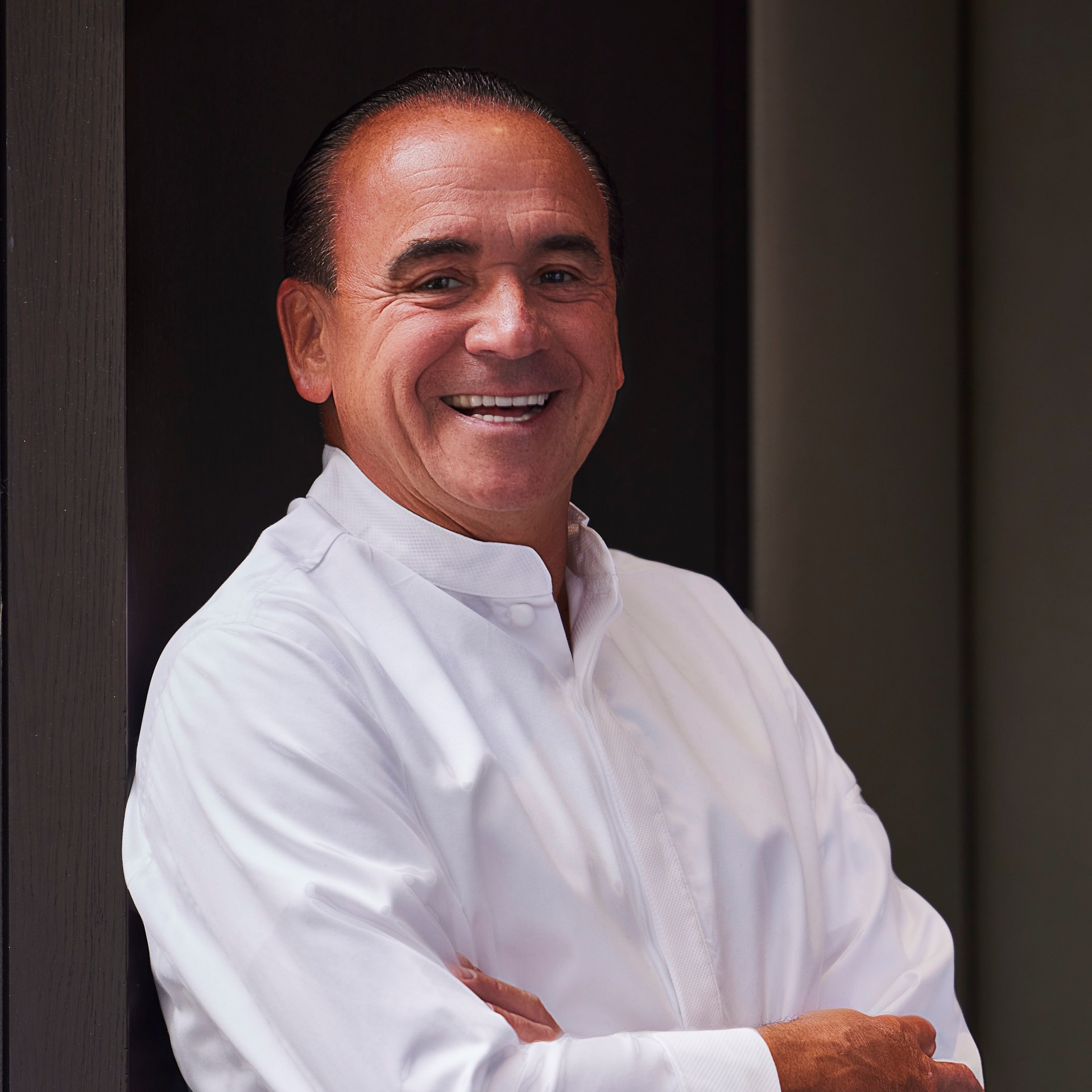 Jean-Georges Vongerichten wearing a white chef's jacket and black pants stands with his arms crossed, leaning against a dark wooden frame in a restaurant setting.