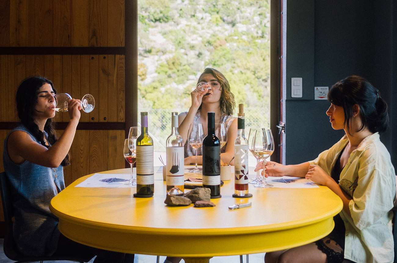 Three women sitting around a yellow table, drinking wine. Four wine bottles on the table.