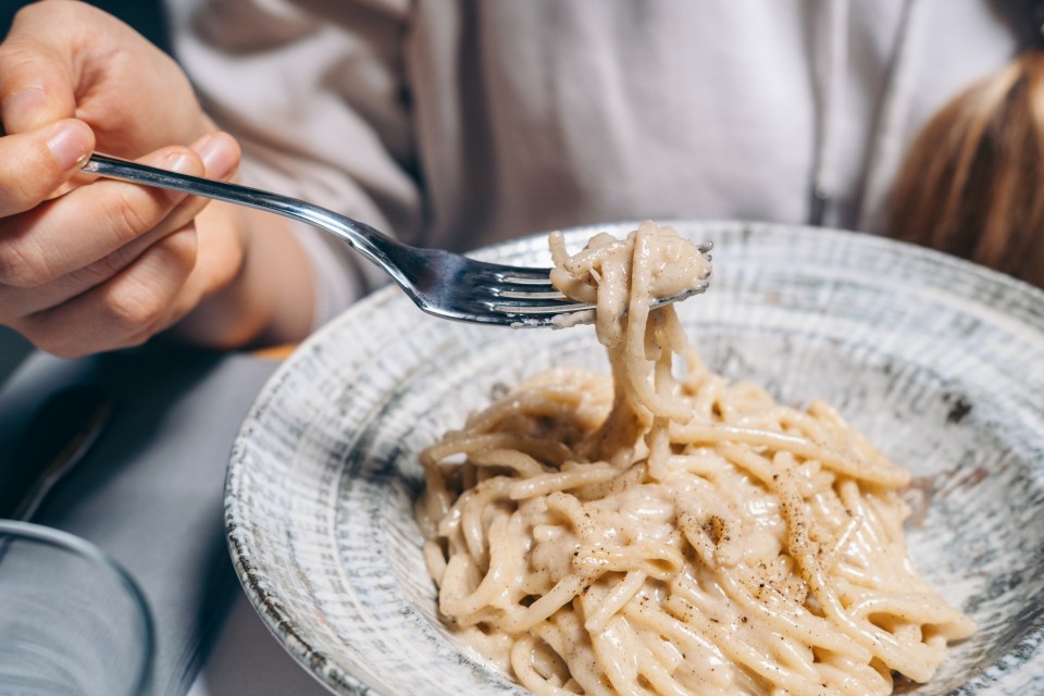 A close-up of a person's hand using a fork to twirl spaghetti covered in a creamy cheese sauce with black pepper from a bowl.