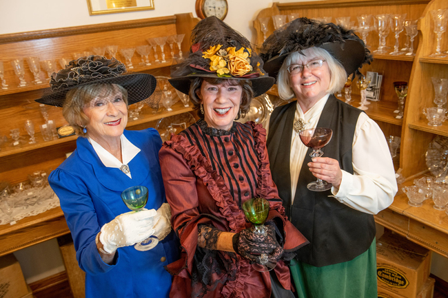 three women in period costumes holding glassware inside the National Museum of Cambridge Glass in Ohio