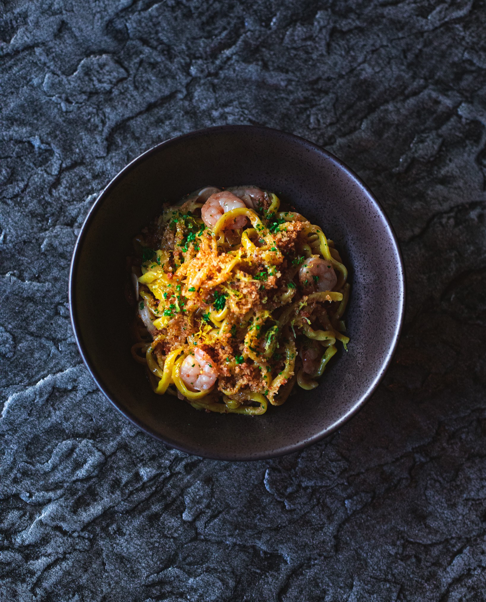 an overhead image of a bowl of shrimp scampi ramen. several shrimp are visible between the thick golden noodles. the noodles are covered with garlic breadcrumbs and parsley. the bowl is dark gray and sits in front of a textured gray background.