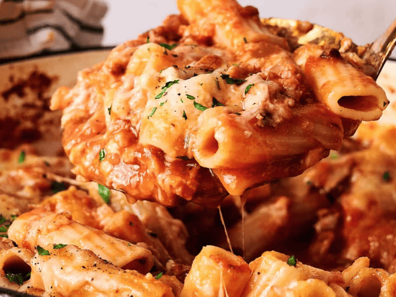 A spoon scoops baked ziti pasta covered in melted cheese and tomato sauce from a casserole dish.