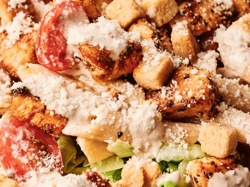 A Caesar salad with grilled chicken, penne pasta, cherry tomatoes, croutons, and shredded cheese, topped with Caesar dressing on a bed of lettuce.
