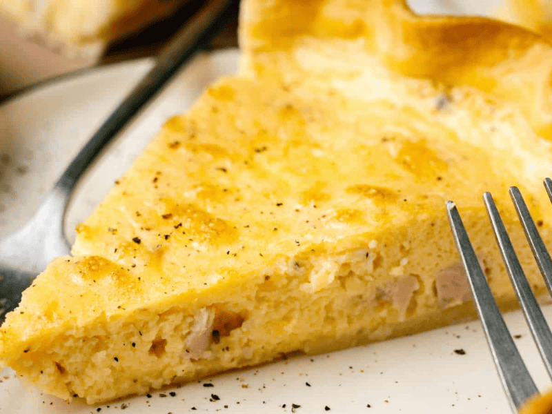 A close-up of a slice of quiche on a plate, with a fork beside it. The quiche has a golden crust and a creamy, egg-filled interior.