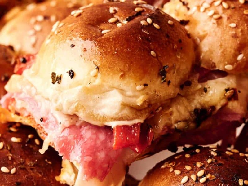 Close-up of sesame seed-topped sliders filled with sliced meat and melted cheese.