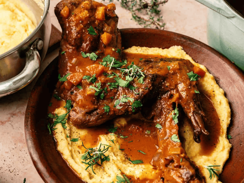 A plate of braised lamb shanks in a tomato-based sauce served over creamy polenta, garnished with fresh herbs.