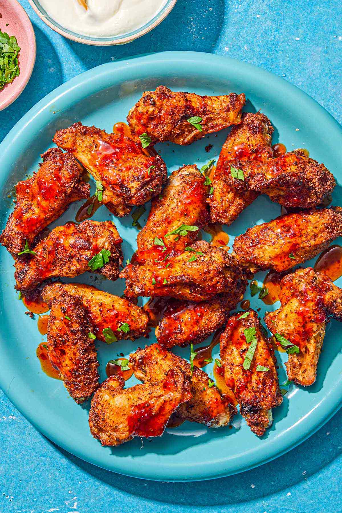 Air fryer chicken wings topped with harissa hot honey and chopped parsley on a platter. Next to this are bowls of creamy feta dressing and chopped parsley.