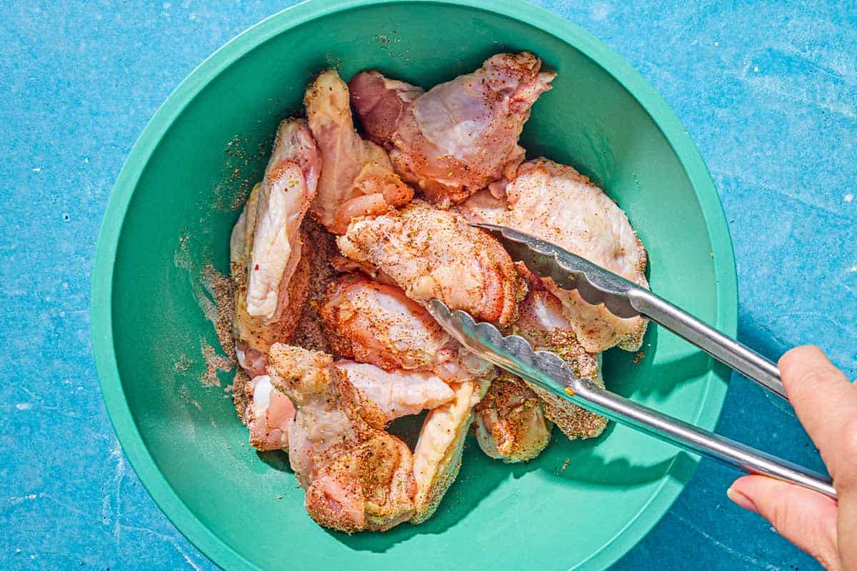 The uncooked chicken wings being mixed with the seasoning in a bowl with a pair of tongs.