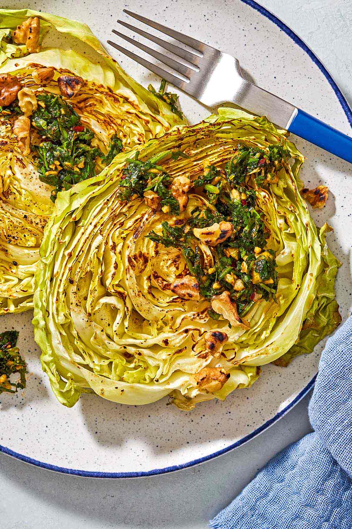 A close up of two cabbage steaks topped with green chermoula and toasted walnuts on a plate with a fork.